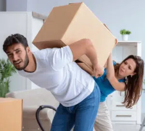 Un ragazzo che cerca di caricarsi sulle spalle uno scatolone pesante mentre la ragazza prova ad aiutarlo
